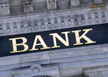 A building facade featuring ornate stone architecture with the word "BANK" prominently displayed in large, gold letters. The background is a dark rectangular panel that contrasts with the light-colored stone, emphasizing its distinguished banking heritage. | ZIIMP