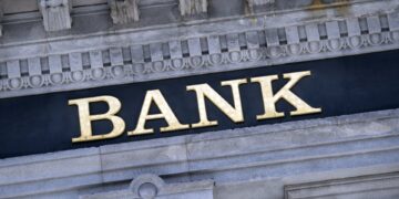 A building facade featuring ornate stone architecture with the word "BANK" prominently displayed in large, gold letters. The background is a dark rectangular panel that contrasts with the light-colored stone, emphasizing its distinguished banking heritage. | ZIIMP