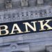 A building facade featuring ornate stone architecture with the word "BANK" prominently displayed in large, gold letters. The background is a dark rectangular panel that contrasts with the light-colored stone, emphasizing its distinguished banking heritage. | ZIIMP