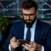 A man in a suit and glasses sits at a desk, reviewing investment strategies on his smartphone. Behind him, large screens display stock market charts. A plant on a shelf to his side adds a touch of greenery to the professional setting. | ZIIMP
