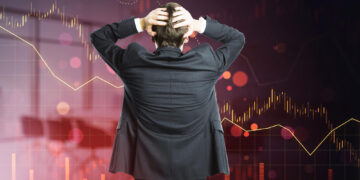 A person in a suit stands with hands on head, facing a large digital screen with declining financial graphs and red trends, symbolizing the challenge of overcoming emotional investing and staying rational in volatile markets. | ZIIMP