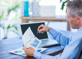 A man in a blue shirt sits at a desk, analyzing printed charts with graphs and bar data. He holds a pen, pointing to a line graph while researching Building your First Investment Portfolio on his laptop with a blank screen open in front of him. | ZIIMP