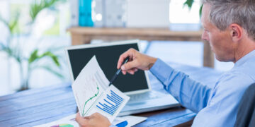 A man in a blue shirt sits at a desk, analyzing printed charts with graphs and bar data. He holds a pen, pointing to a line graph while researching Building your First Investment Portfolio on his laptop with a blank screen open in front of him. | ZIIMP