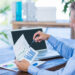 A man in a blue shirt sits at a desk, analyzing printed charts with graphs and bar data. He holds a pen, pointing to a line graph while researching Building your First Investment Portfolio on his laptop with a blank screen open in front of him. | ZIIMP
