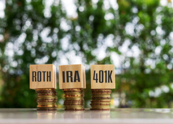 Three stacks of coins topped with wooden blocks labeled "ROTH," "IRA," and "401K" highlight the differences between IRA vs Roth IRA vs 401k, set on a reflective surface against a blurred green natural background. | ZIIMP
