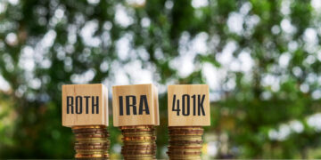 Three stacks of coins topped with wooden blocks labeled "ROTH," "IRA," and "401K" highlight the differences between IRA vs Roth IRA vs 401k, set on a reflective surface against a blurred green natural background. | ZIIMP