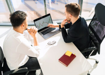Two men sitting at a desk in an office, looking at a laptop screen displaying stock market data. A notebook labeled "What is Investing," along with a pen and eyeglasses, are on the white desk near them. | ZIIMP