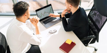 Two men sitting at a desk in an office, looking at a laptop screen displaying stock market data. A notebook labeled "What is Investing," along with a pen and eyeglasses, are on the white desk near them. | ZIIMP