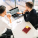 Two men sitting at a desk in an office, looking at a laptop screen displaying stock market data. A notebook labeled "What is Investing," along with a pen and eyeglasses, are on the white desk near them. | ZIIMP