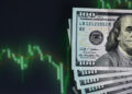 A stack of 100-dollar bills lies in the lower right corner beside a digital screen showing a green, declining stock market graph on a dark background, highlighting the potential benefits of Dollar-Cost Averaging during downturns. | ZIIMP