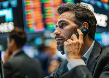 A man in a suit talks on a phone headset at a busy stock exchange, surrounded by colorful digital stock boards—an environment perfect for those eager to learn how to start options trading. | ZIIMP