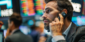 A man in a suit talks on a phone headset at a busy stock exchange, surrounded by colorful digital stock boards—an environment perfect for those eager to learn how to start options trading. | ZIIMP