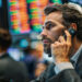 A man in a suit talks on a phone headset at a busy stock exchange, surrounded by colorful digital stock boards—an environment perfect for those eager to learn how to start options trading. | ZIIMP