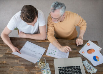 Two people sit at a wooden table with financial charts, a laptop, cash, coins in a jar, and documents, discussing How to Set Smart Financial Goals: A Beginner's Guide to Investing. They are viewed from above, focused on the papers in front of them. | ZIIMP