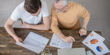 Two people sit at a wooden table with financial charts, a laptop, cash, coins in a jar, and documents, discussing How to Set Smart Financial Goals: A Beginner's Guide to Investing. They are viewed from above, focused on the papers in front of them. | ZIIMP