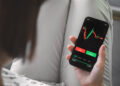 A person holding a smartphone with a stock trading app open, displaying a candlestick chart and buy and sell buttons—showing how easy it is to read the stock market. The person's legs are crossed, dressed in light-colored pants. | ZIIMP