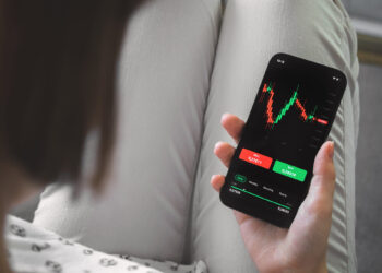 A person holding a smartphone with a stock trading app open, displaying a candlestick chart and buy and sell buttons—showing how easy it is to read the stock market. The person's legs are crossed, dressed in light-colored pants. | ZIIMP