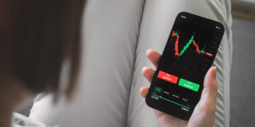 A person holding a smartphone with a stock trading app open, displaying a candlestick chart and buy and sell buttons—showing how easy it is to read the stock market. The person's legs are crossed, dressed in light-colored pants. | ZIIMP