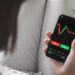 A person holding a smartphone with a stock trading app open, displaying a candlestick chart and buy and sell buttons—showing how easy it is to read the stock market. The person's legs are crossed, dressed in light-colored pants. | ZIIMP