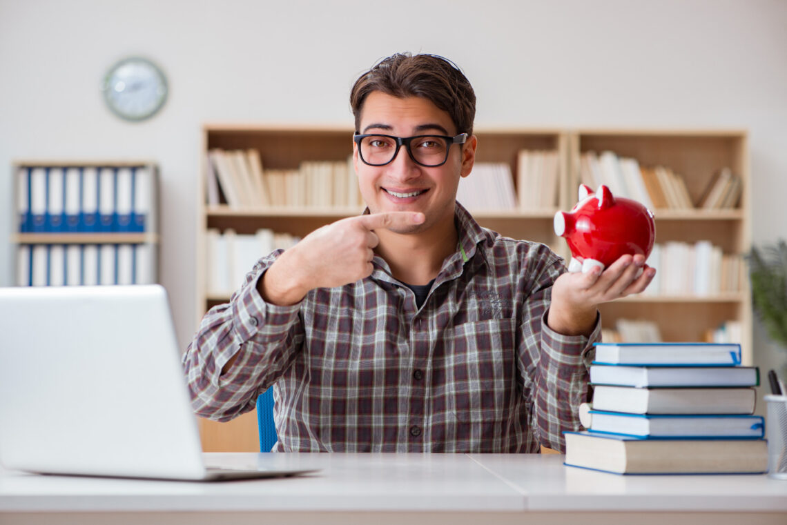 A smiling man wearing glasses sits at a desk with a laptop and books, holding and pointing at a red piggy bank. Bookshelves and a clock are in the background. | ZIIMP