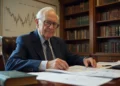 Warren Buffett, sits at a desk with financial documents in an office lined with bookshelves. A stock market Value Investing Secrets chart hangs on the wall behind him as light streams through a window to his left. | ZIIMP